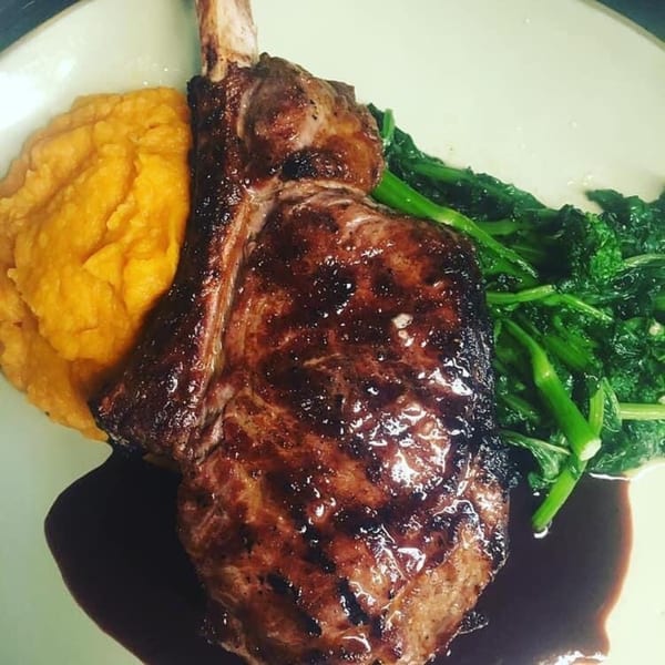 Grilled bone-in steak with mashed sweet potatoes, green vegetables, and a dark sauce.